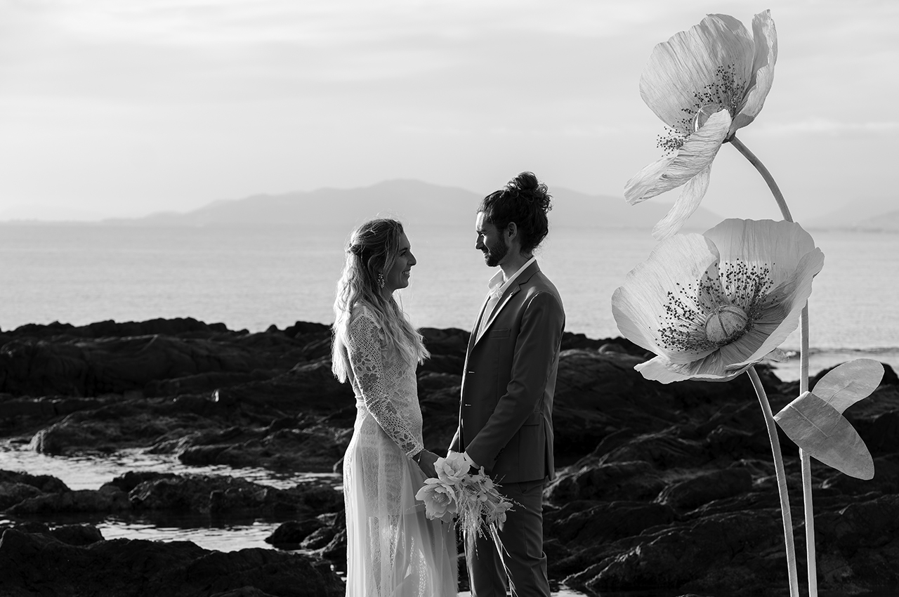 Un couple de mariés se tient face à face devant deux fleurs géantes blanches en papier créées par Étoile Vagabonde, au bord de l’eau